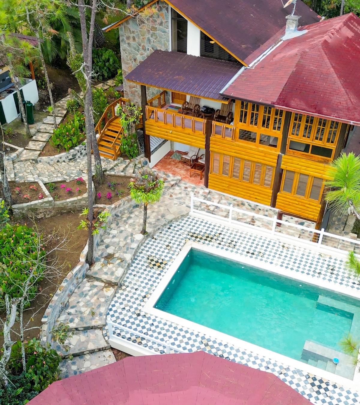 An aerial view reveals a villa built with natural stone and wood, featuring a red-tiled roof. A swimming pool is positioned beside a stone pathway adorned with greenery, leading to a spacious outdoor deck furnished with wooden seating.