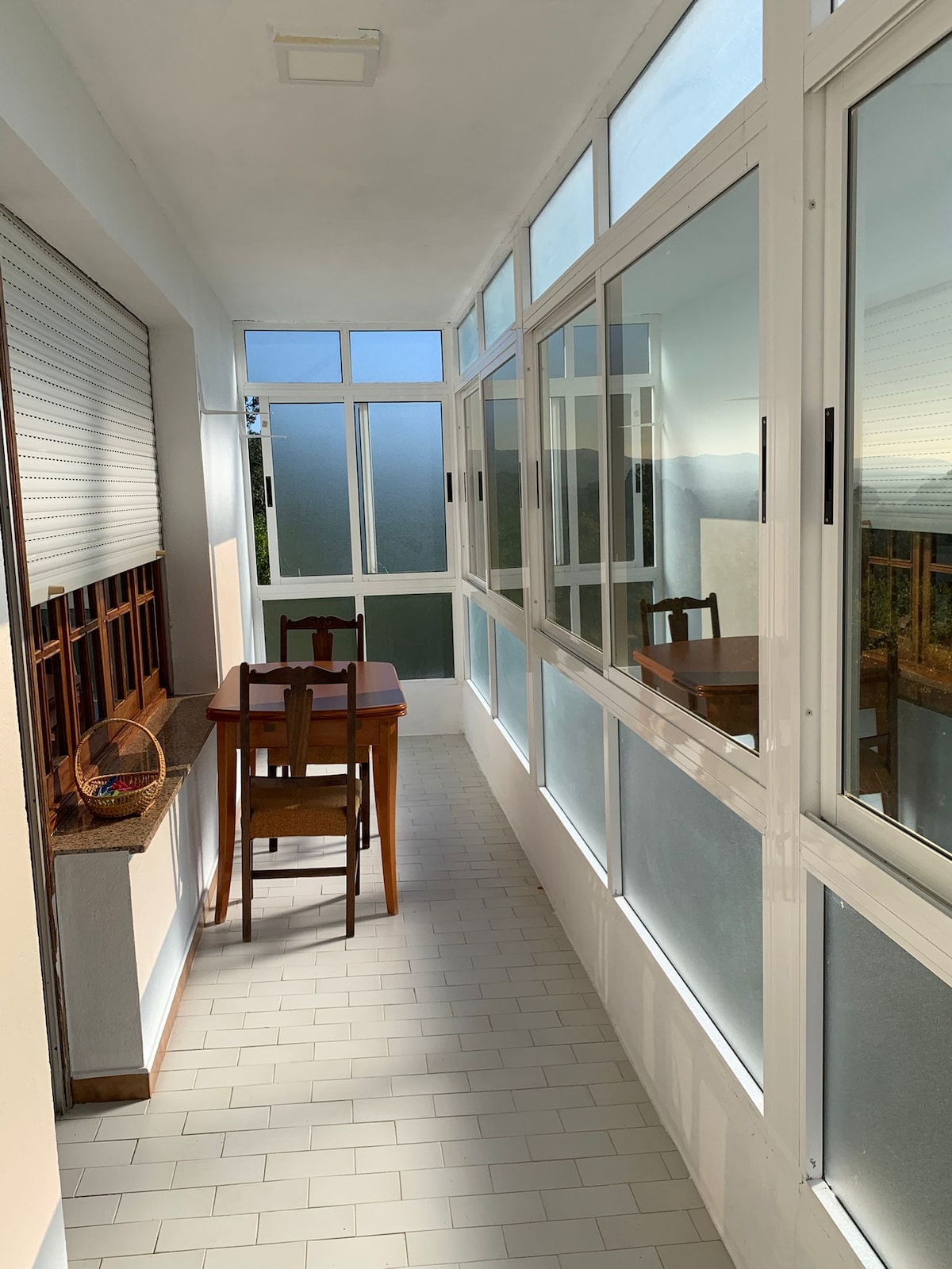 A sunlit enclosed balcony is visible, featuring multiple large windows with frosted glass. A small wooden table and a chair are arranged against the wall. The flooring is tiled in a light color, contributing to the airy feel of the space.