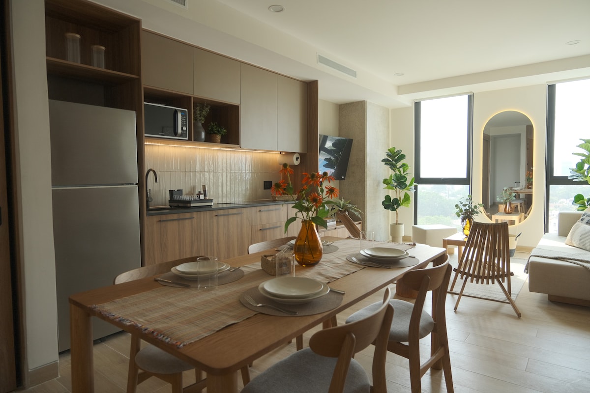 An open-plan living space features a modern kitchen with stainless steel appliances and wooden cabinetry. A dining table is set with neutral dishware, surrounded by light-colored chairs. A comfortable lounge area is adorned with potted plants, and large windows allow natural light to fill the room.