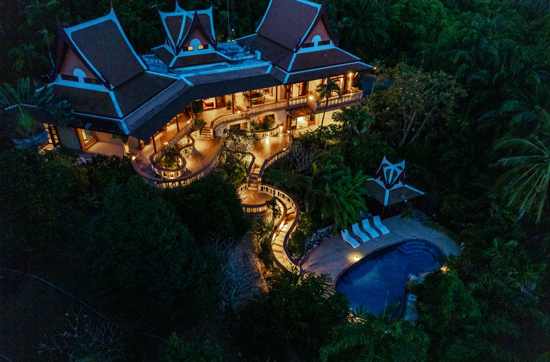 The villa is beautifully illuminated at night, showcasing its elegant architecture against a lush tropical backdrop. A private saltwater pool is visible surrounded by palm trees and outdoor seating areas, offering a serene atmosphere in the hillside retreat.