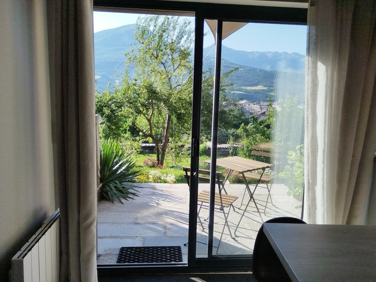 A sliding glass door provides views of the garden area, framed by trees and plants. Outside, two folding chairs and a small table are visible on a stone patio. The distant mountains can be seen under a clear sky, enhancing the serene environment.