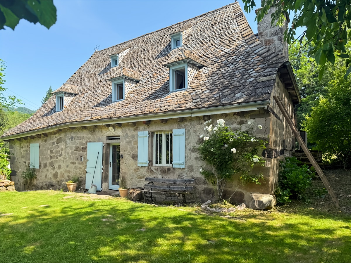 The exterior of a charming stone house is presented, featuring a distinctive slate roof and light blue shutters. Lush greenery surrounds the property, with a well-maintained lawn and a wooden bench positioned near the entrance. The inviting garden adds to the tranquil atmosphere.