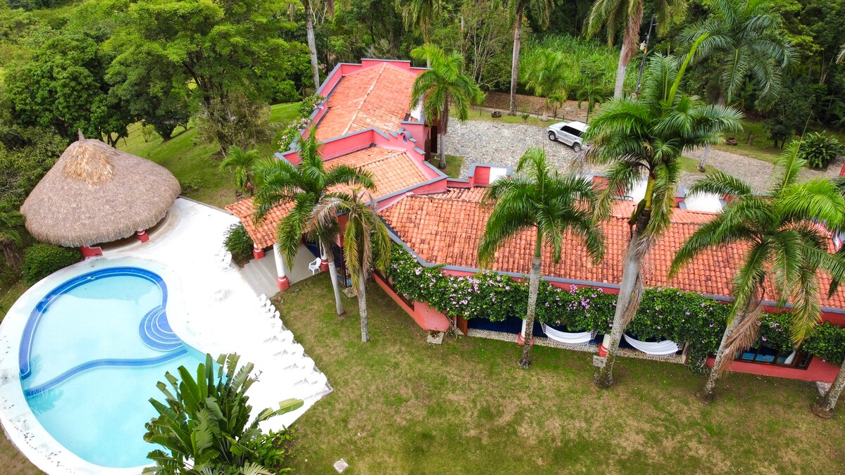 The property is viewed from above, revealing a spacious layout with a vibrant red-tiled roof. An inviting pool curves beside a thatched-roof gazebo, surrounded by lush greenery and palm trees. The natural landscape complements the colorful architecture, emphasizing a tranquil and tropical atmosphere.
