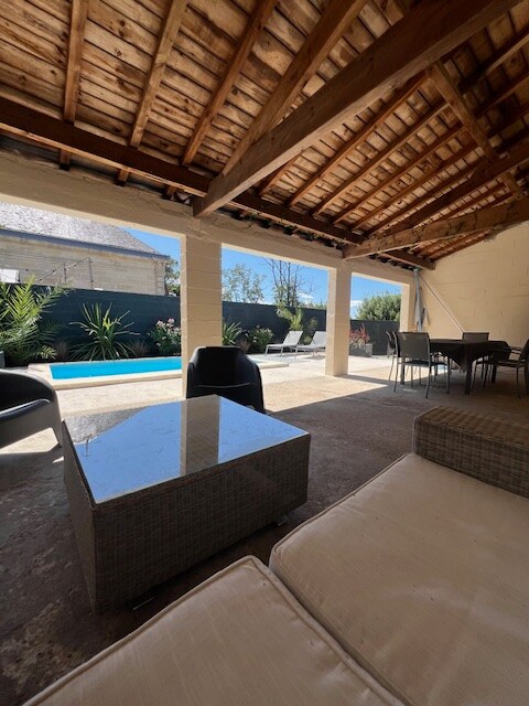 An outdoor patio area features a comfortable seating arrangement with a low table. In the background, a private pool is visible, surrounded by greenery. The overhead wooden beams add a rustic charm to the open space.