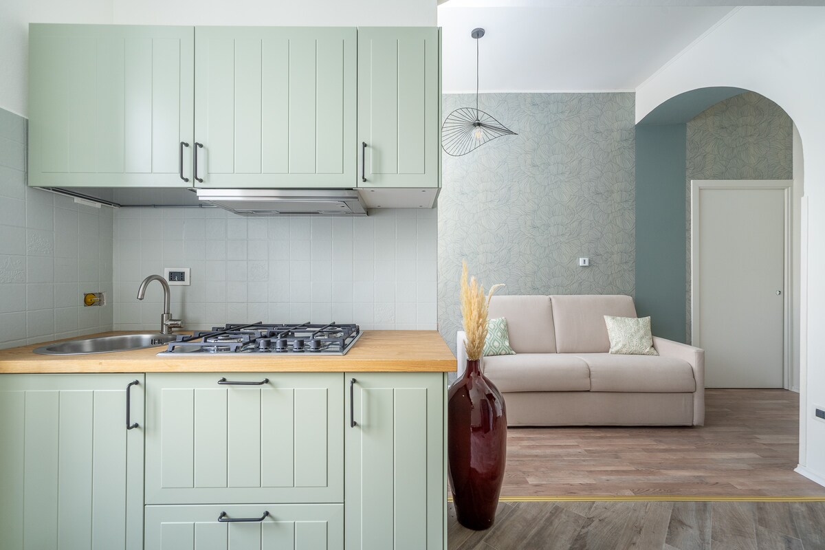 A modern kitchen area features soft green cabinetry and a wooden countertop. A stovetop is integrated into the cabinetry. Adjacent, a light-colored sofa is placed against a decorative wall, creating a welcoming space. A floor vase with dried grasses adds a touch of elegance.