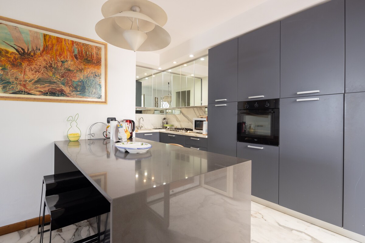 The modern kitchen features a sleek black island with a glossy surface, complemented by stylish black chairs. Stainless-steel appliances, including an oven and microwave, are set within dark cabinetry. A decorative artwork hangs on the wall, contributing to the kitchen's contemporary ambiance.