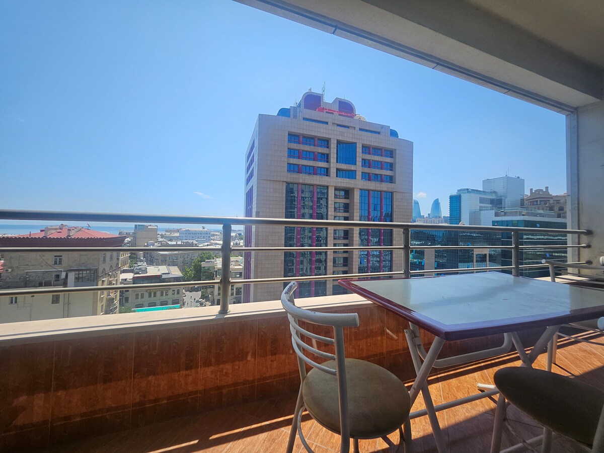 A spacious balcony is presented, offering panoramic views of the skyline and cityscape. A modern table with four chairs is visible, inviting relaxation in the sunlight. The scene features a clear blue sky and buildings, contributing to a vibrant urban atmosphere.