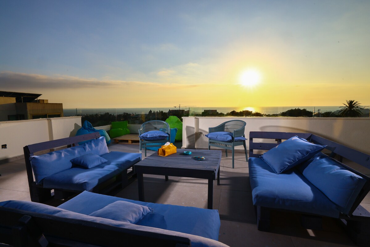 A spacious rooftop terrace is equipped with comfortable seating, featuring blue cushions on wooden frames. A low table is positioned in the center, and the background showcases a sunset over the sea, with soft light illuminating the scene.
