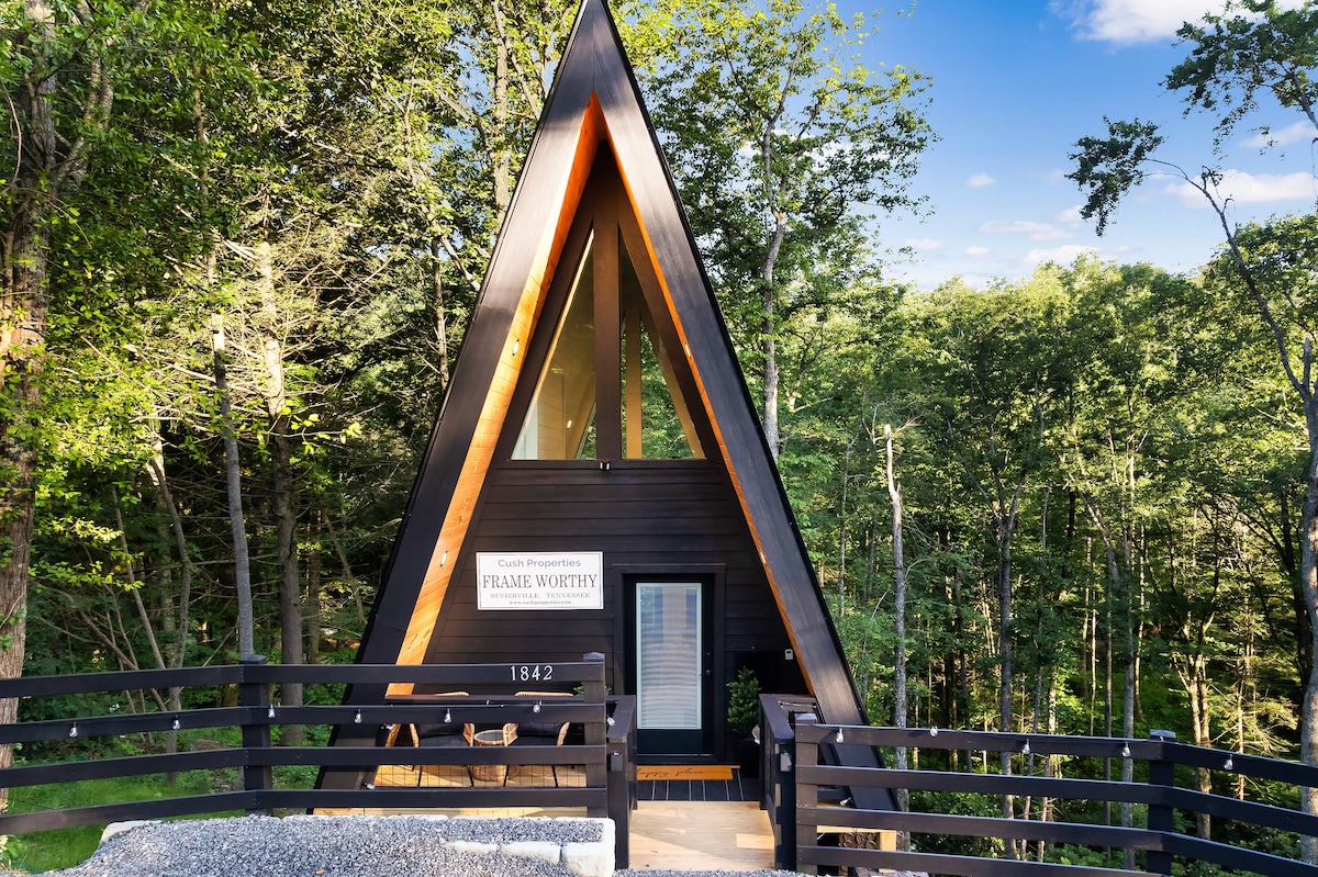The A-frame cabin, framed by lush greenery, features a striking triangular design with wooden accents. A welcoming entrance is highlighted by a simple sign, and a wraparound deck allows for easy access to the outdoors. The scene is bathed in natural light.