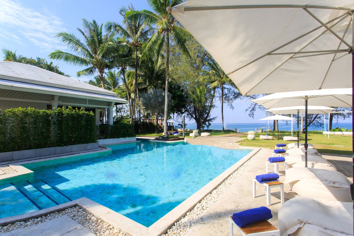 A serene pool area is surrounded by tropical trees and comfortable lounge chairs with blue cushions. Large umbrellas provide shade, and the inviting pool reflects the clear sky, with the beach visible in the background, creating a calm and relaxing atmosphere.