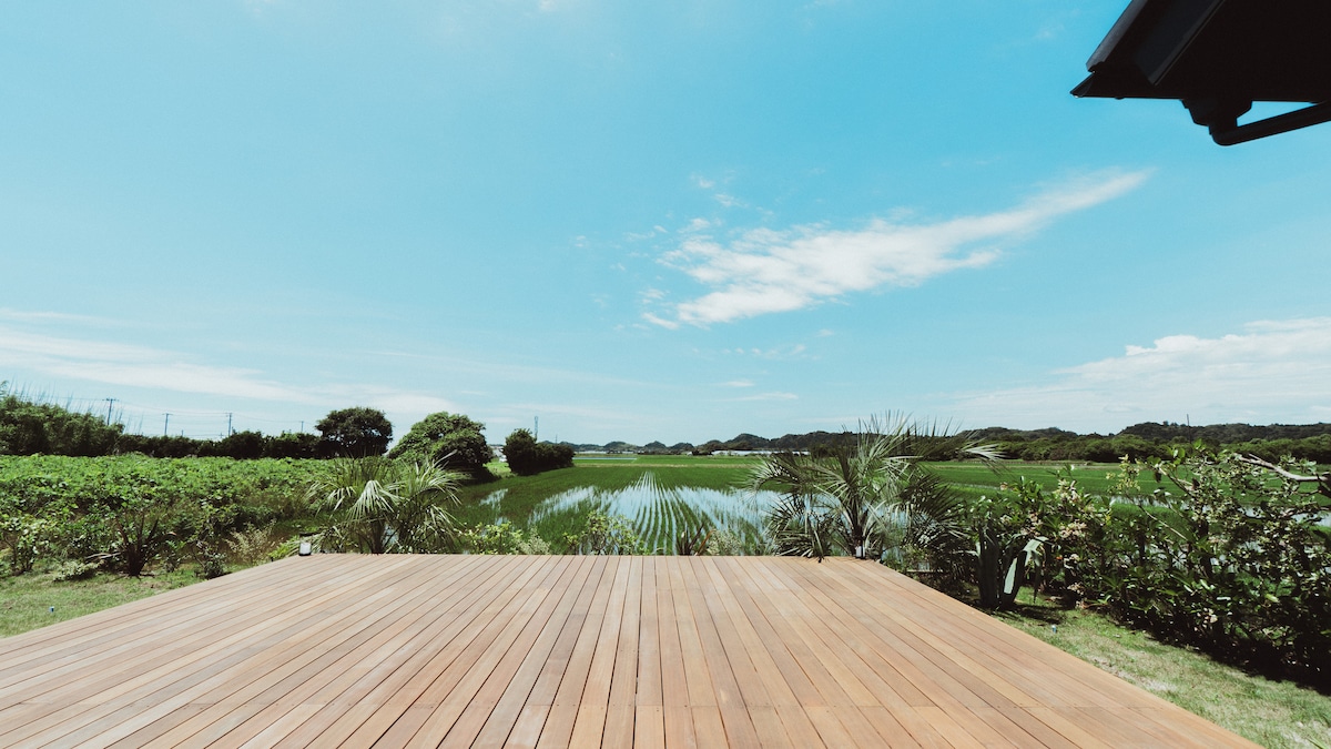 A spacious wooden deck extends out towards expansive rice fields, creating a seamless connection with nature. Lush greenery frames the view, while the clear blue sky enhances the serene atmosphere of the retreat. The deck invites moments of relaxation and reflection.