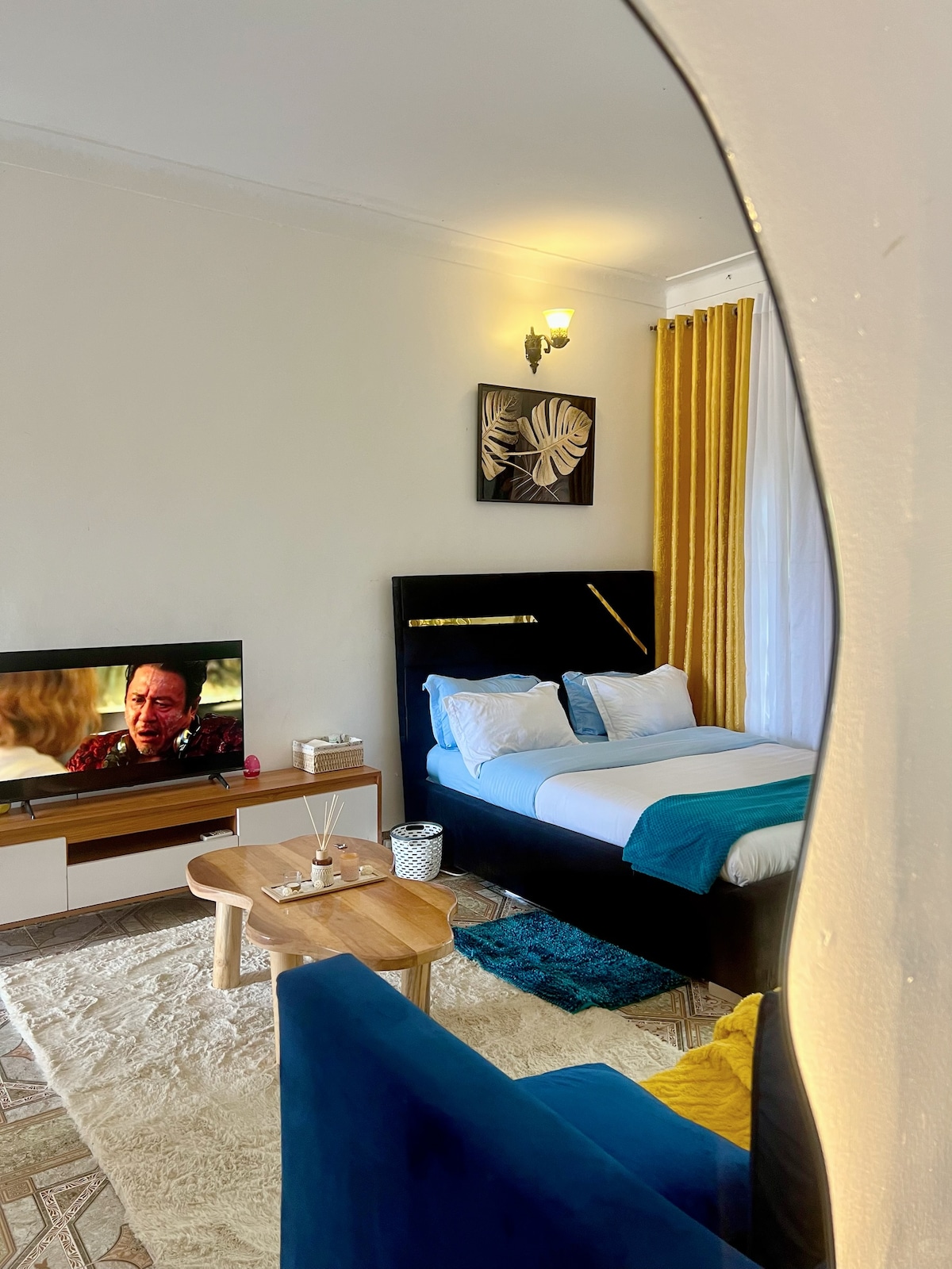 A room features a sleek black bed with white and blue bedding, complemented by vibrant yellow curtains. A wooden coffee table sits in front of a blue accent chair, and a television is positioned on a modern media console. Natural light fills the space.