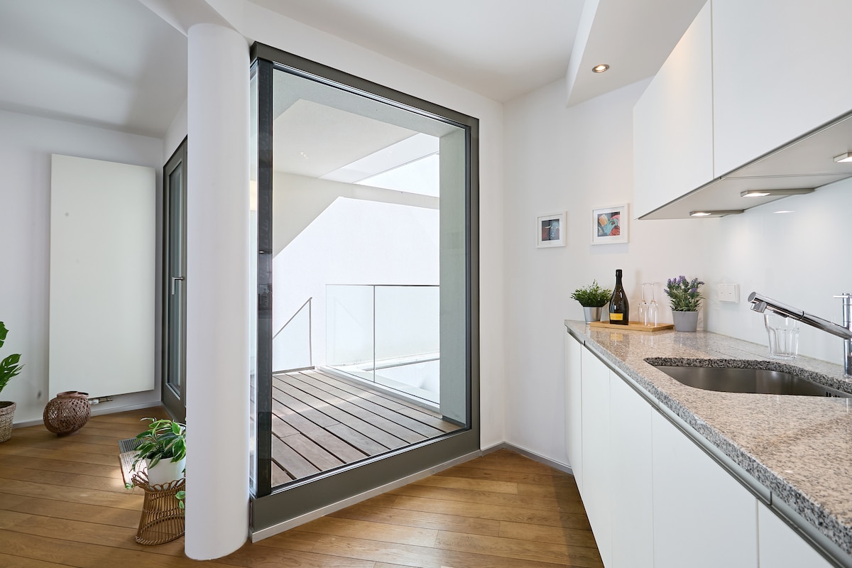 A modern kitchen area is presented, featuring sleek countertops and minimalistic cabinetry. Large glass doors offer a view of a wooden terrace, allowing natural light to fill the space. Potted plants and decorative items enhance the ambiance, creating a fresh and inviting atmosphere.