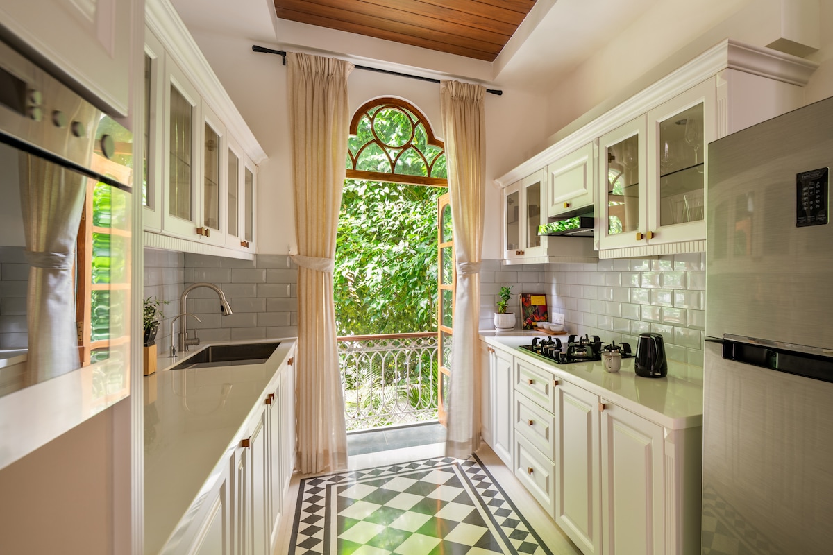 The fully equipped kitchen features elegant cabinetry and modern appliances. Natural light flows in through large windows, enhancing the fresh atmosphere. A small balcony is visible, providing a view of the greenery outside. The patterned floor adds a touch of charm to the space.