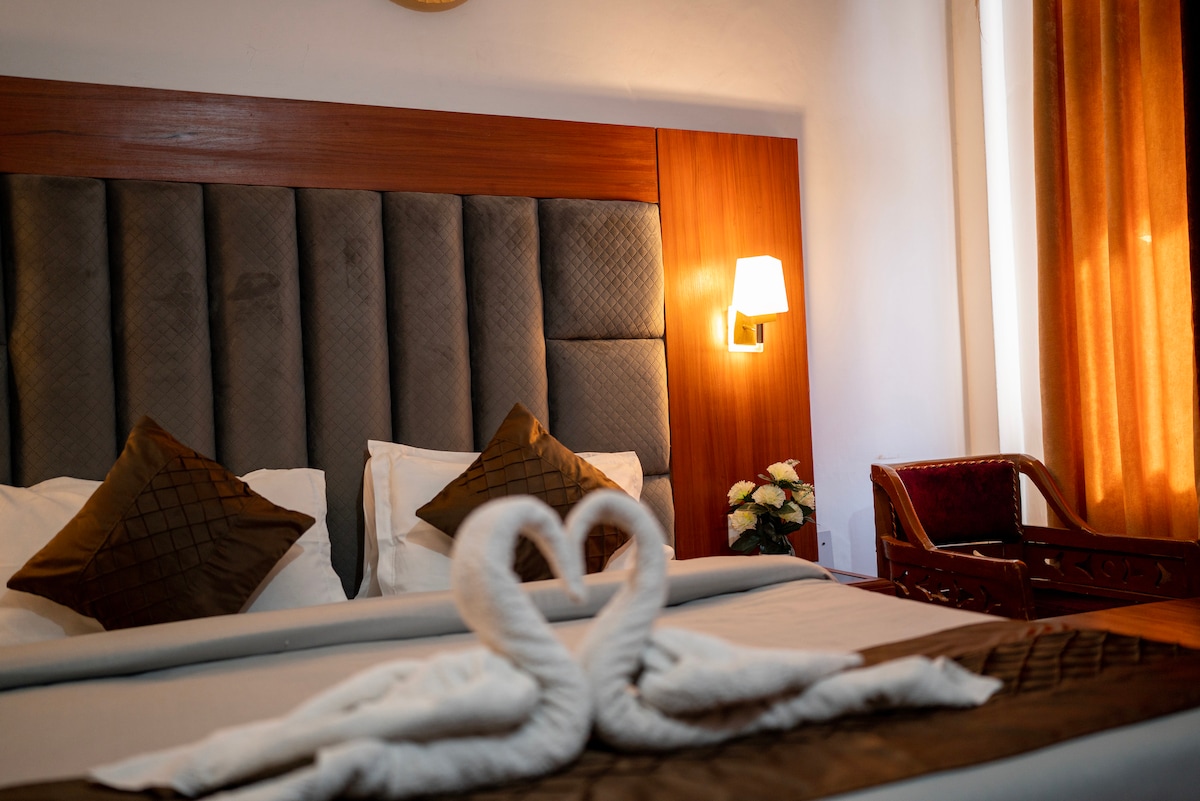 A cozy sleeping area features a stylish headboard with a textured design. Two swan-shaped towels are arranged on the plush bedding. Soft, warm lighting is provided by a wall-mounted lamp, and a small, wooden chair is positioned near the window, which is dressed in light curtains.