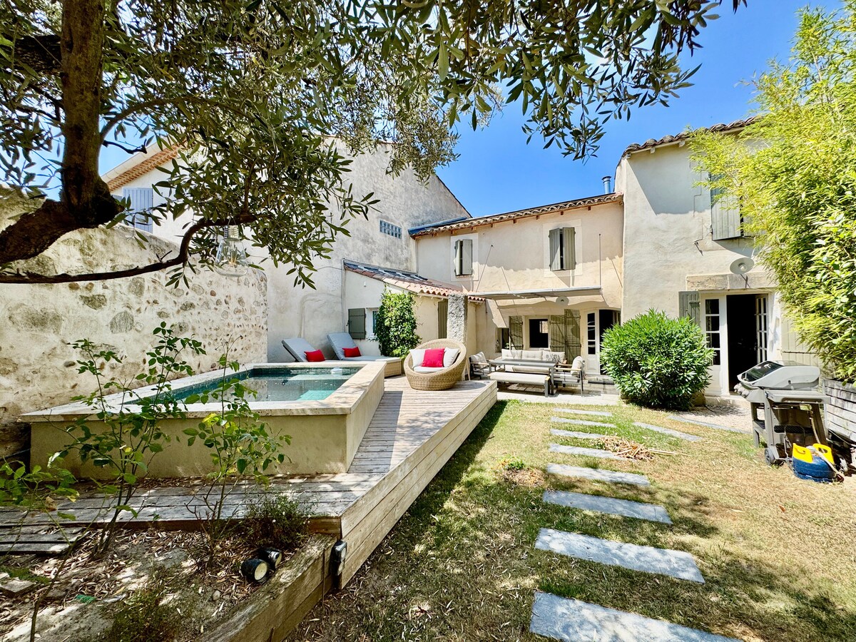 The exterior of the home features a well-maintained garden with a wooden deck surrounding a small heated pool. Comfortable seating areas are arranged with cushions, and a pathway of stone slabs leads through the greenery, complementing the relaxed outdoor ambiance.
