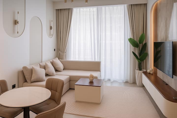 Sala de estar luminosa y elegante con gran ventanal, cortinas opacas, sofá esquinero beige, mesa de centro de madera lacada, mesa de comedor redonda con sillas, suelo claro y decoración de pared depurada. Ambiente acogedor, moderno y cálido.