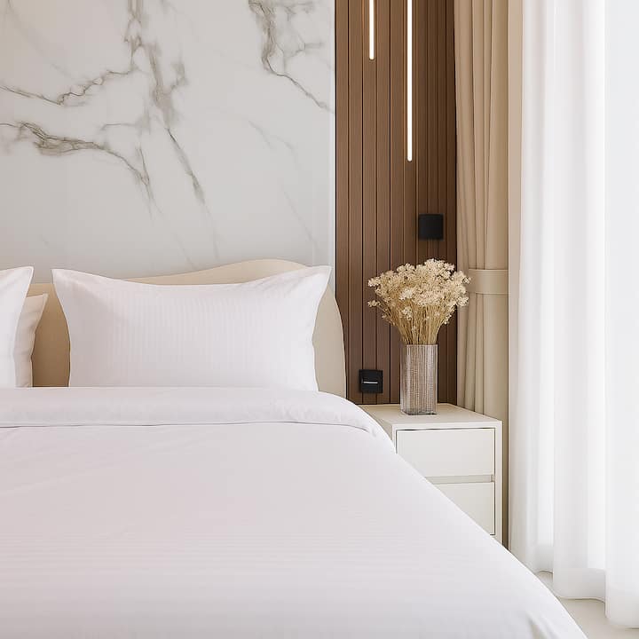 Elegante cabecero con panel de mármol y madera nervada, iluminación suspendida de diseño, mesita de noche blanca y decoración floral suave. Ambiente relajante y refinado.