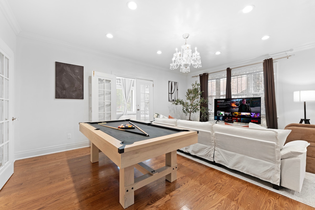 A spacious living room is furnished with a large sectional sofa adorned with light-colored upholstery and plush pillows. A well-lit chandelier hangs above a modern pool table situated in the center, while a 65-inch Smart TV is positioned against the wall, providing entertainment options.