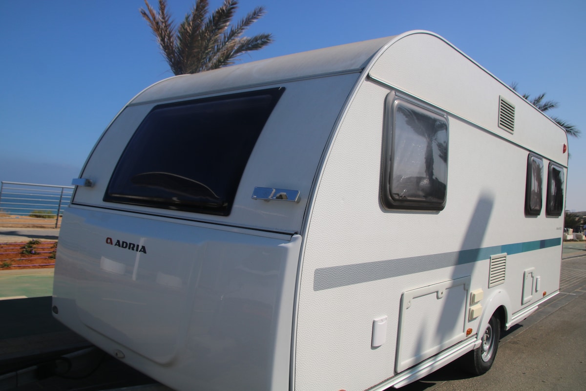 A well-maintained caravan is shown parked along a scenic route with palm trees in the background. The exterior features a clean white design with light blue accents and large windows, providing an inviting and spacious appearance.