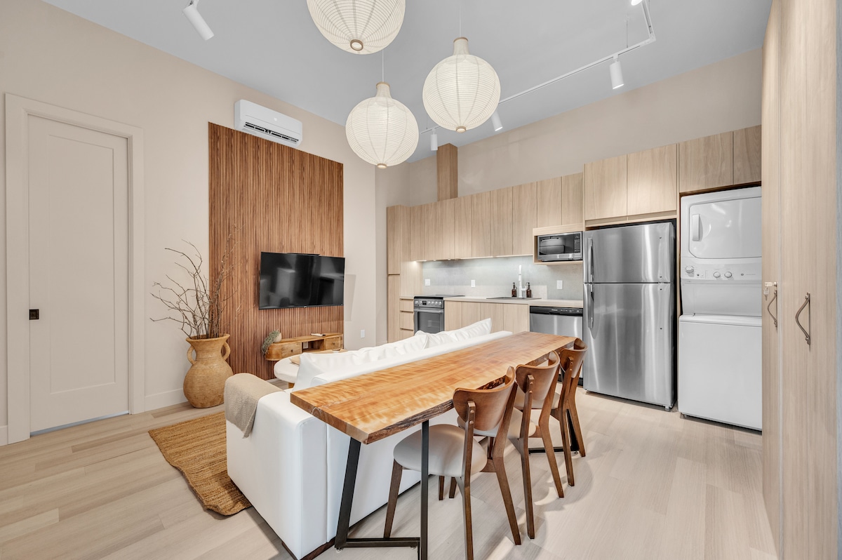 An open-concept kitchen and dining area is displayed, featuring a wooden dining table with four chairs. Modern appliances, including a refrigerator and a stacked washer and dryer, are integrated into the cabinetry. Soft lighting is provided by hanging paper lanterns, creating a calm atmosphere.