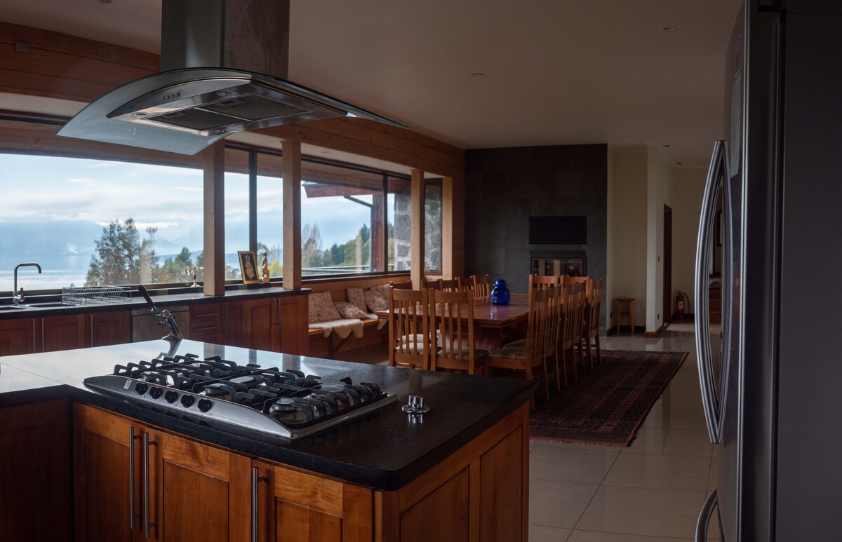 A spacious kitchen featuring an island counter with a gas stove, surrounded by wooden cabinetry. Large windows provide a view of the outdoors and fill the area with natural light. A dining table with multiple seats is visible in the background, along with a cozy seating area.
