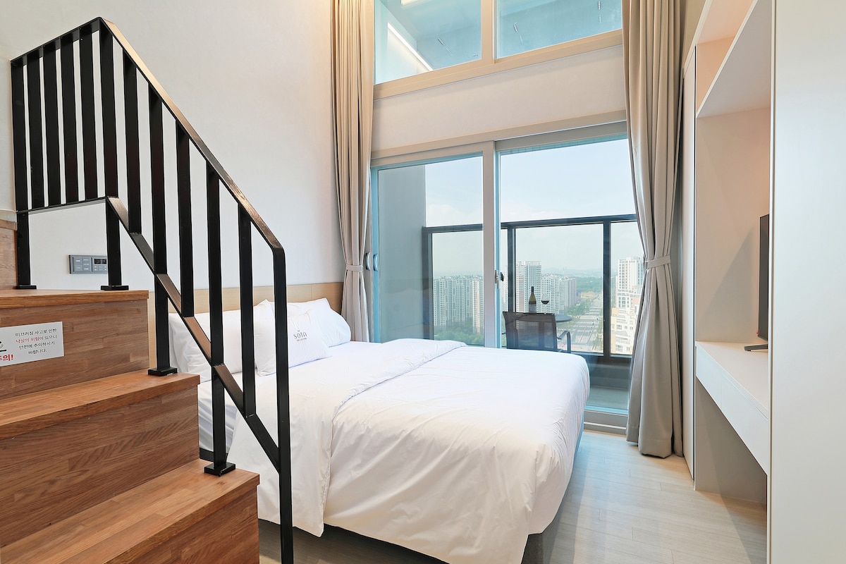 A well-lit bedroom features a spacious bed with crisp white linens. Large windows provide a clear view of the outside, while a modern staircase leads to an upper level. Neutral tones and minimalist furnishings contribute to a clean and open atmosphere.