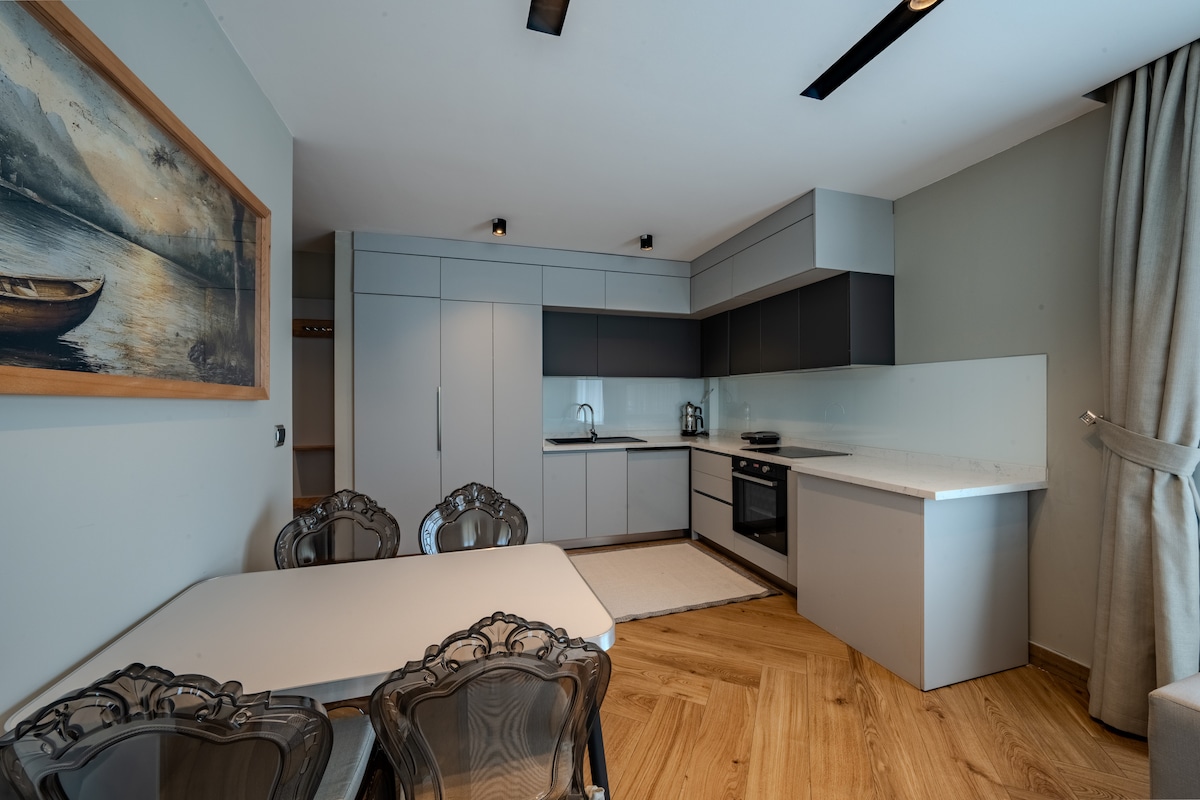 A modern kitchen area features sleek cabinetry in contrasting colors, a built-in oven, and various kitchen amenities. A dining table with stylish chairs is positioned nearby, complemented by light-colored flooring. Natural light enhances the space through a nearby window partially covered by elegant curtains.