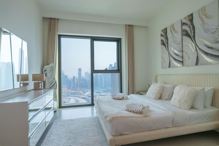 Chumba cha kulala chenye mwangaza chenye kitanda cha kifahari, mapambo maridadi na madirisha ya sakafu hadi dari yanayoangalia anga ya jiji.