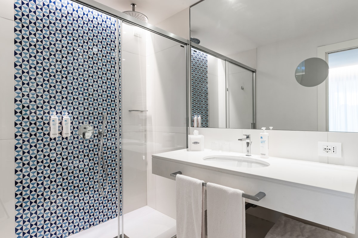 A modern bathroom featuring a walk-in shower enclosed by glass panels. A stylish tiled wall with a geometric pattern adds visual interest. A white sink and countertop are complemented by neatly arranged toiletries. Soft towels are displayed on a holder beneath the sink.
