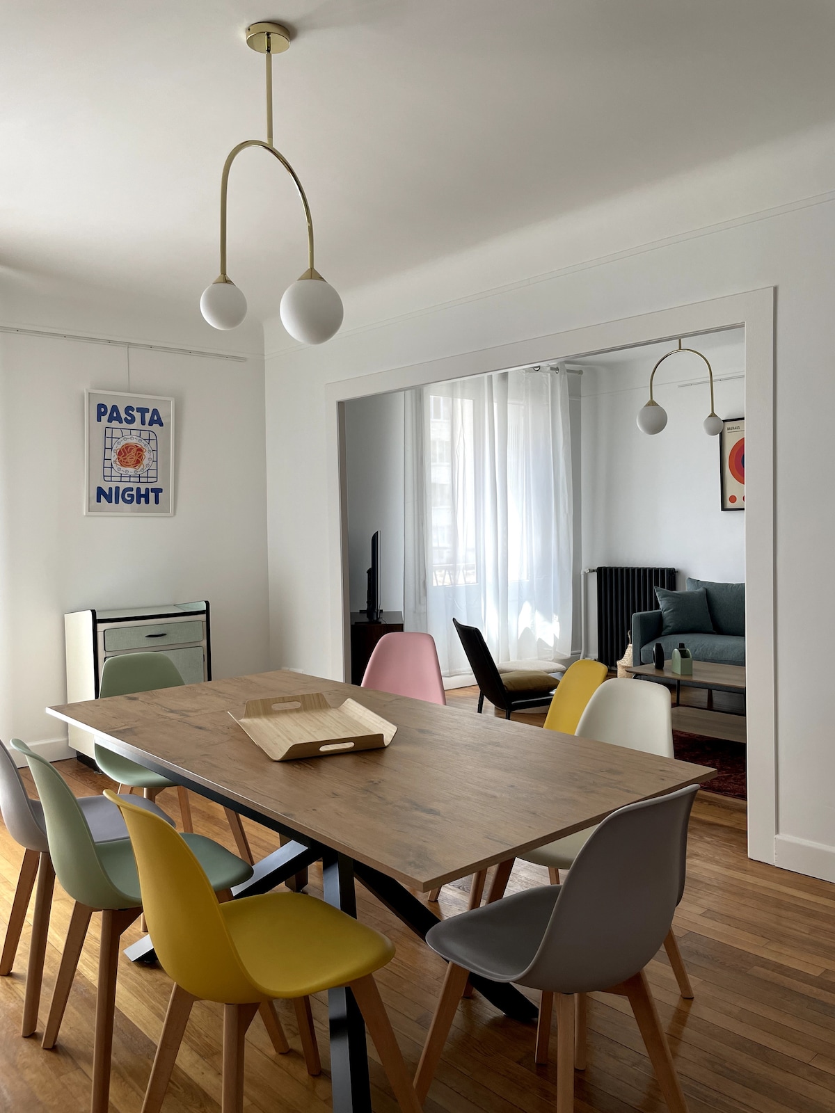 A dining area features a wooden table surrounded by colorful chairs in shades of yellow, gray, and pink. A stylish light fixture hangs overhead, and natural light is let in through sheer curtains. A cozy living space is visible in the background.