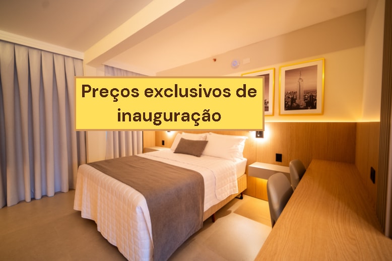 The image displays a bedroom featuring a spacious bed with neutral bedding. Two framed pictures are placed above a modern wooden desk, which is accompanied by two chairs. Soft drapes frame a window that allows light to enter the room.