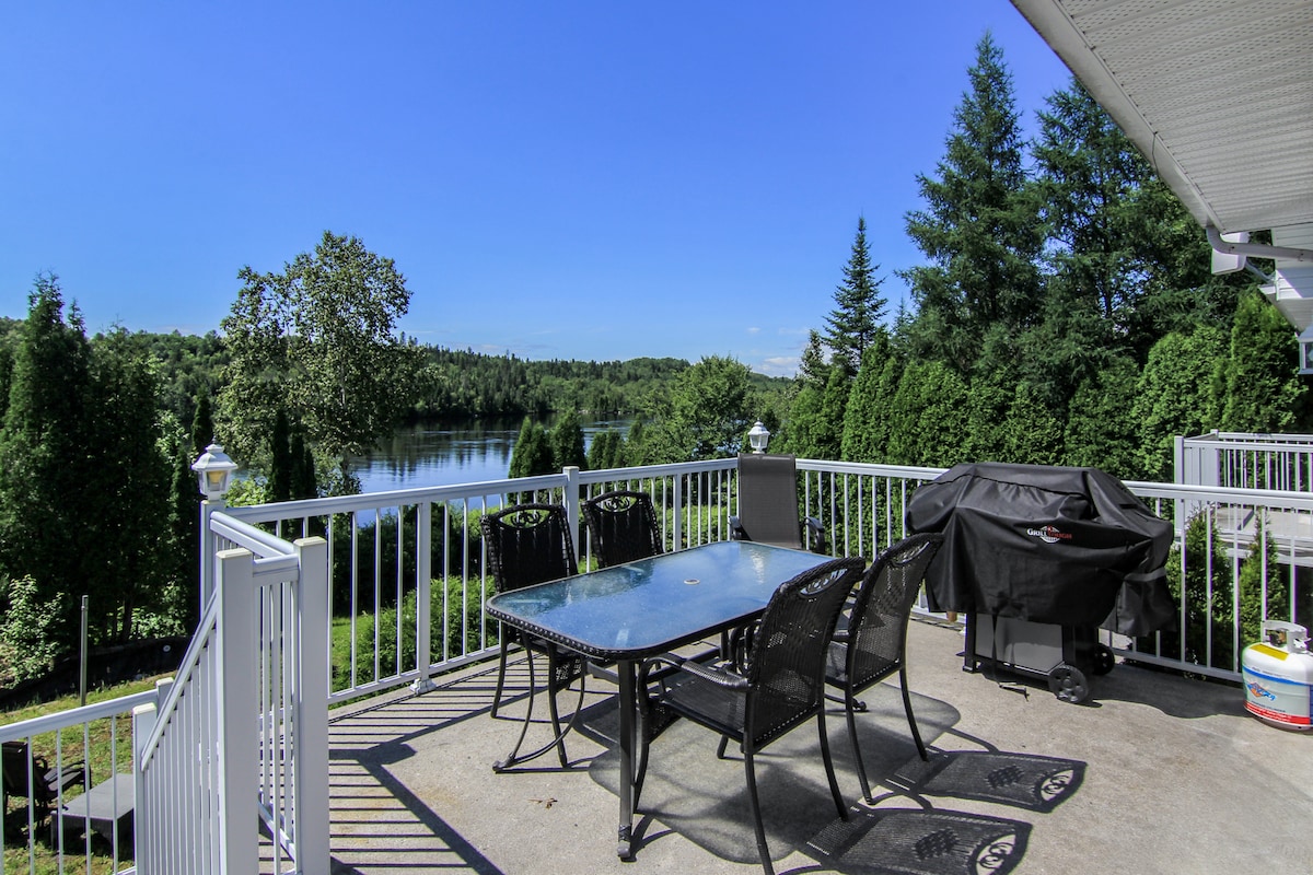 A spacious outdoor patio is equipped with a large table surrounded by several chairs, with a grill nearby. Lush greenery and trees frame the scenic view of the water, offering a serene backdrop under a clear blue sky.