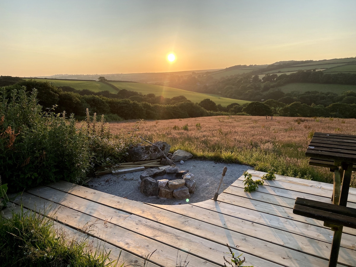 A wooden decking area is showcased with a small fire pit surrounded by stones. Beyond, expansive fields and rolling hills are visible, illuminated by a warm sunset, creating a serene natural environment.
