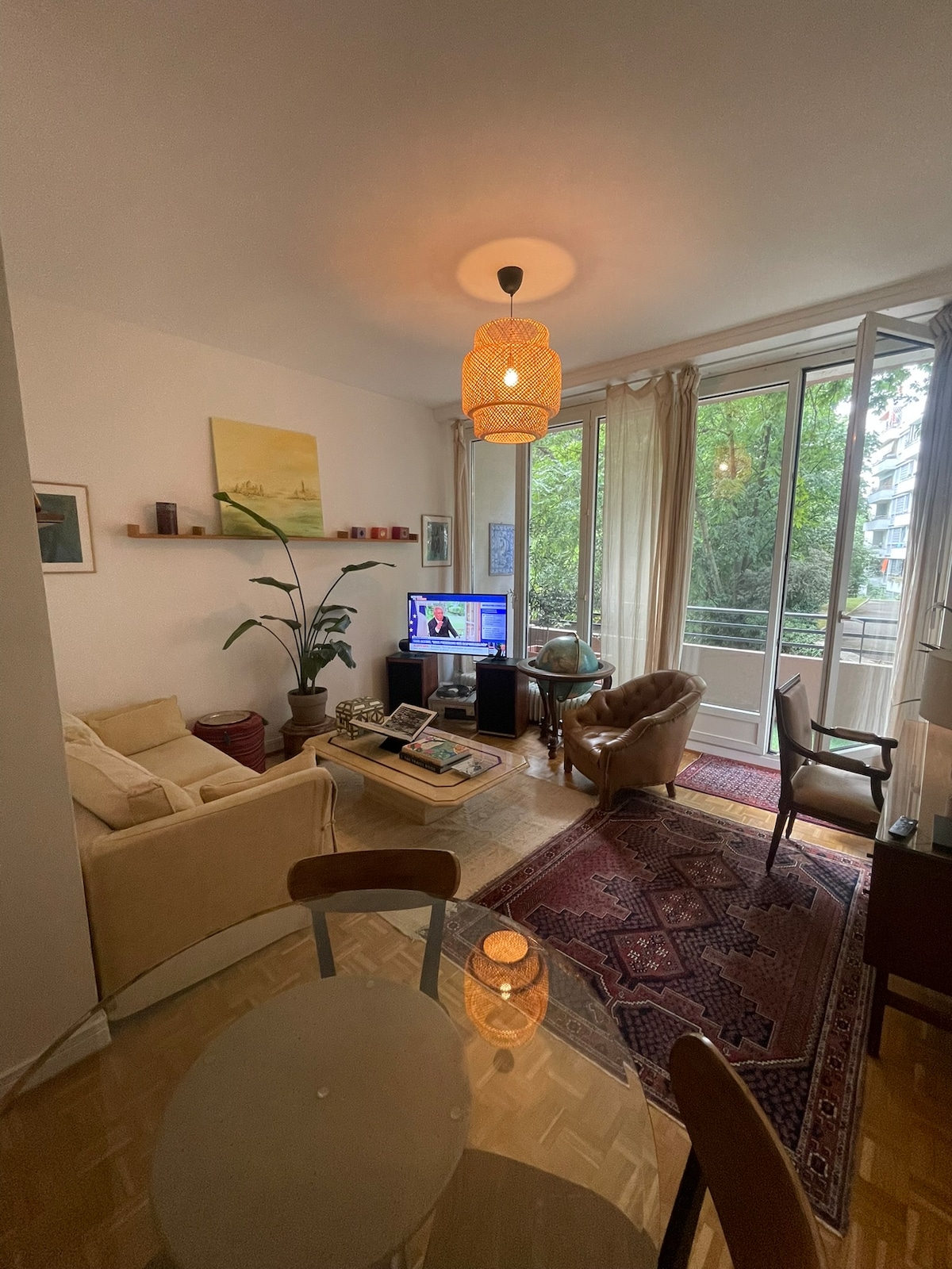 The living area features a comfortable seating arrangement with a light-colored sofa and a brown armchair situated around a decorative area rug. A TV is positioned near large windows, allowing natural light to illuminate the space, which overlooks greenery outside.