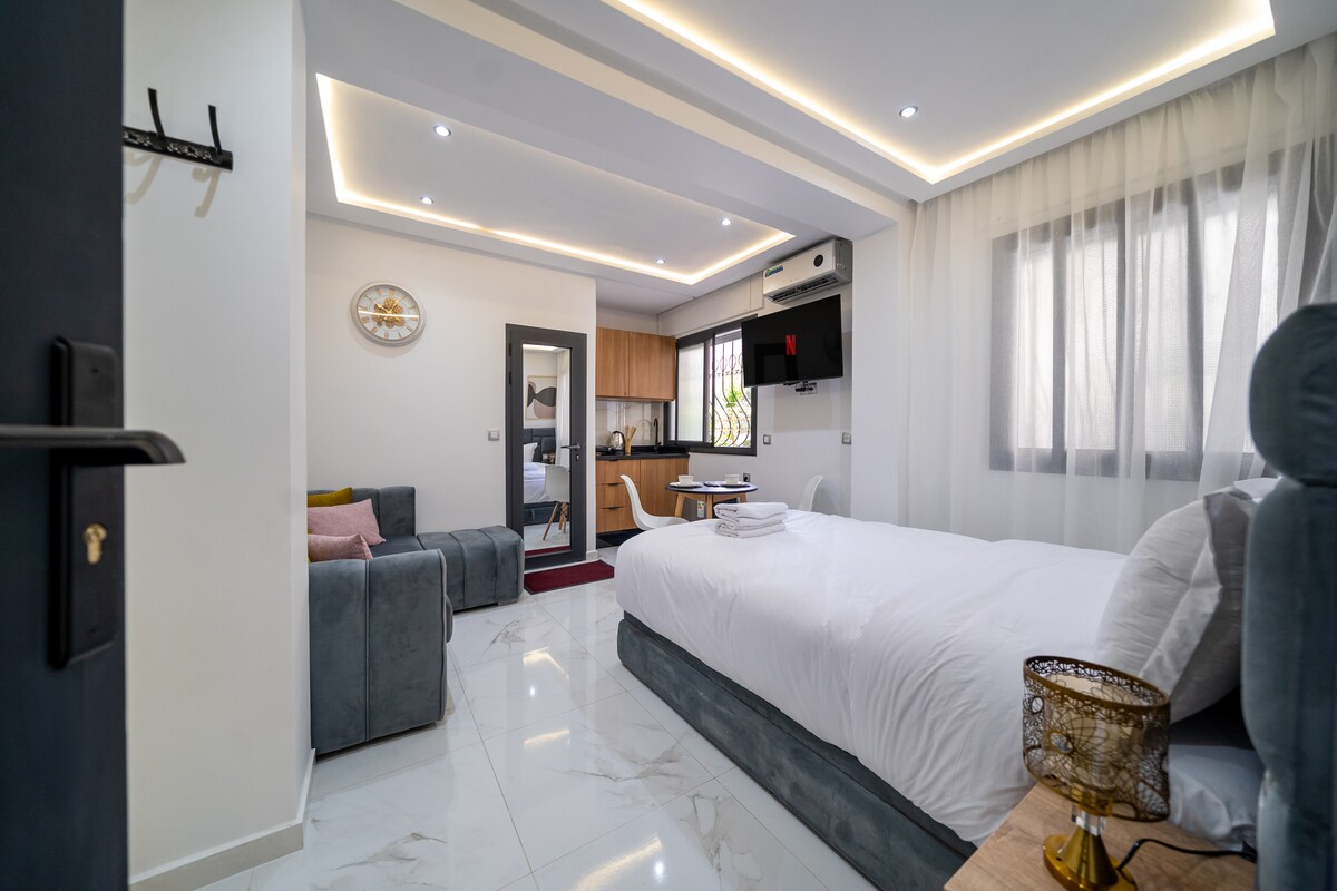 A modern studio room is presented, featuring a spacious bed covered with a light comforter. A gray sofa is visible in the corner, complemented by a small side table. Natural light flows through sheer curtains, while a kitchenette and television are subtly located in the background.