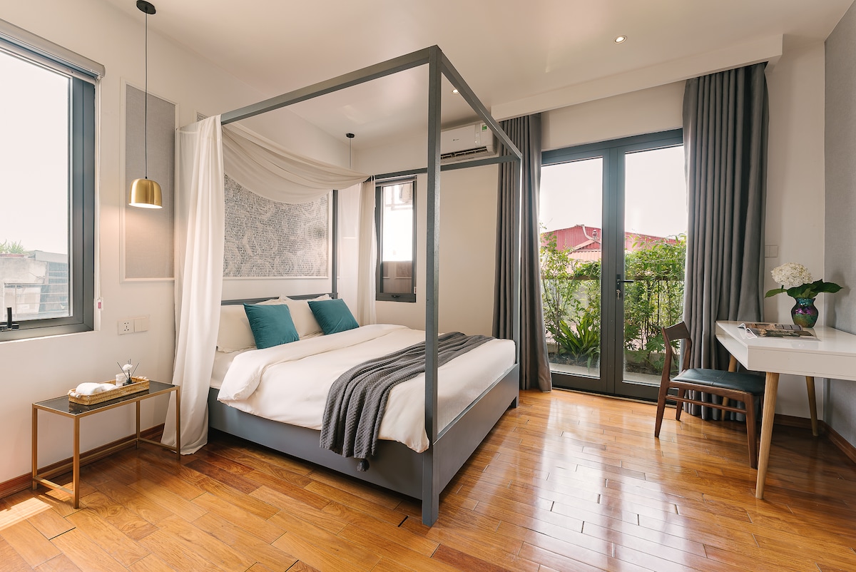 A well-appointed bedroom features a four-poster bed with a draped canopy and two accent pillows. Natural light filters through large glass doors, leading to an outdoor view. A small desk and chair complement the warm wooden flooring and decor.