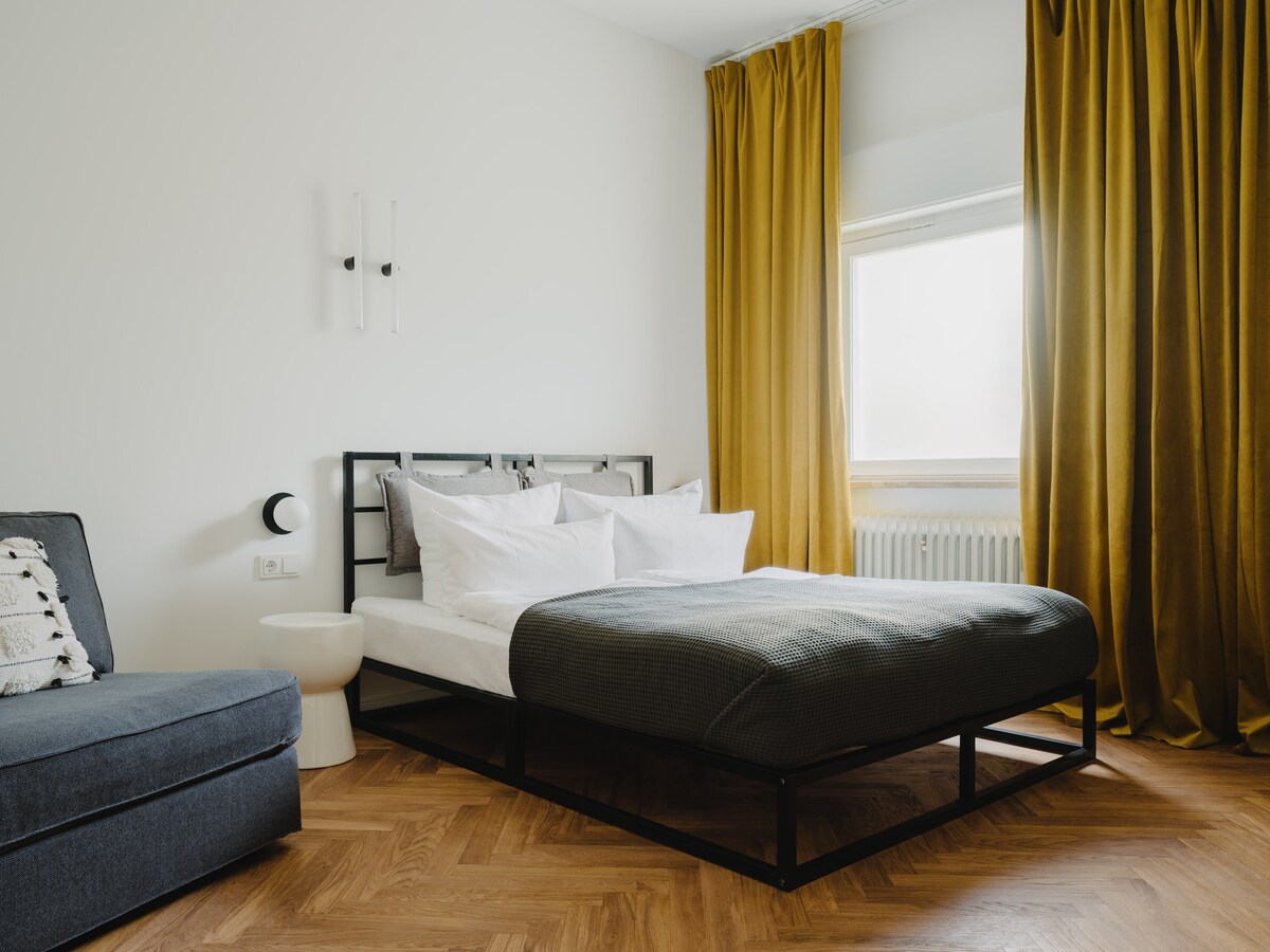 ภาพรวมของห้องนอนที่สองพร้อมเตียงคู่ขนาดควีนไซส์และโซฟาเบดสำหรับคนเดียว ผ้าม่านสีเหลืองอ่อนช่วยให้ห้องดูอบอุ่น 