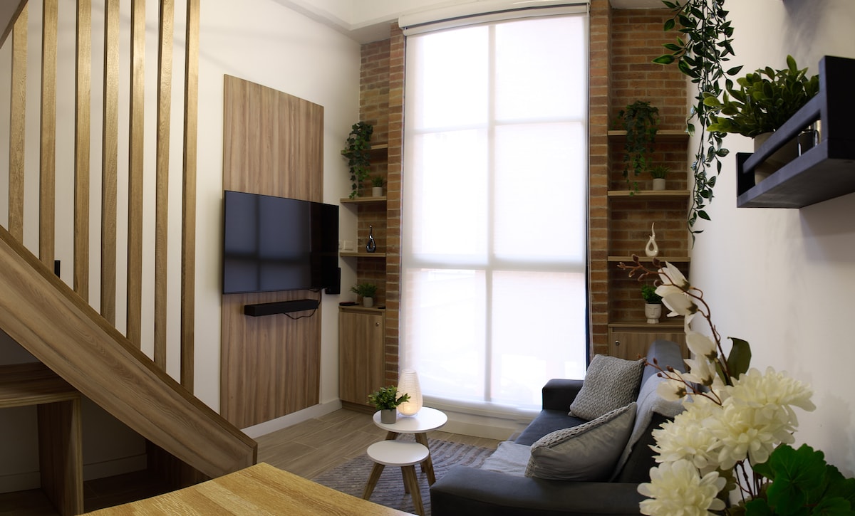 The living area features a cozy sofa positioned next to a wooden coffee table. A large window allows natural light to fill the space, framed by soft sheer curtains. Natural elements are incorporated with plants on shelves and a decorative floral arrangement nearby.