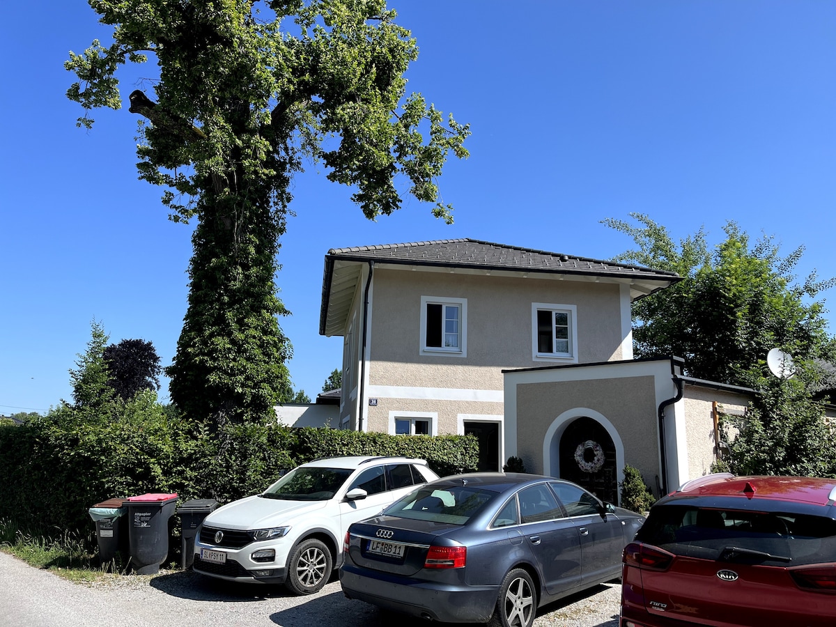 A two-story house is surrounded by greenery and features a mix of light and dark exterior elements. Below, parked cars are visible along the driveway. The structure is complemented by a well-maintained garden area, enhancing the overall outdoor setting.