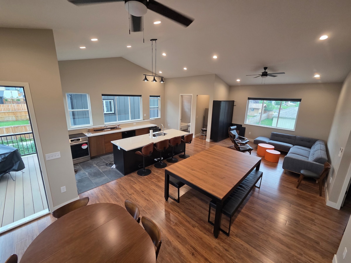 An open-concept living area features a spacious dining table, modern seating arrangements, and a large kitchen island. Natural light fills the room through multiple windows. A sliding door leads to an outdoor space, enhancing connectivity with nature.