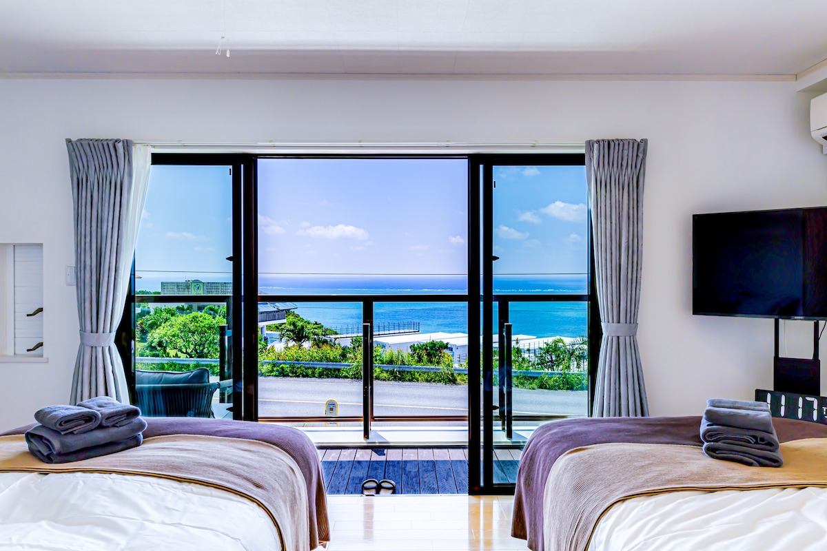 A serene bedroom features two spacious beds with light blankets, positioned for a scenic view through large glass doors. The ocean is visible in the distance, complemented by a bright sky. A television is mounted on the wall opposite the beds, enhancing the room's comfort.