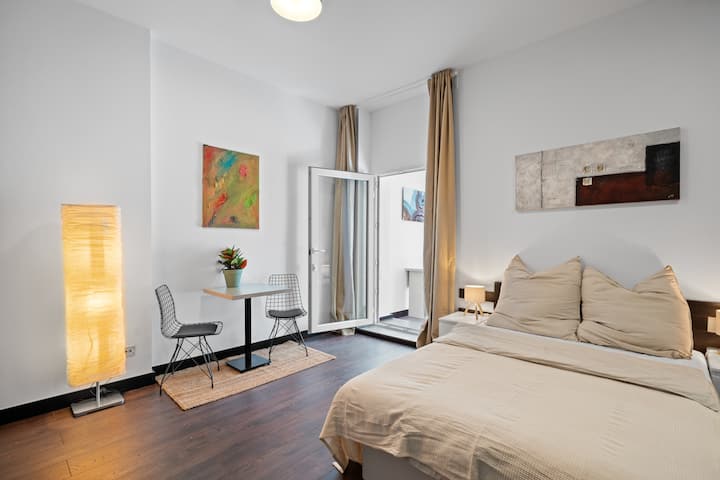 Helles Wohn-Schlafzimmer mit Doppelbett, Esstisch und Wintergartenzugang zum ruhigen Innenhof. Ideal zum Entspannen – stilvoll eingerichtet mit modernen Akzenten.