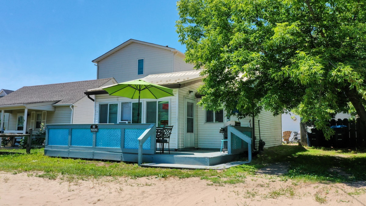 A cozy cottage is set just steps from the beach, featuring a private deck with outdoor seating under a large green umbrella. Lush greenery surrounds the property, and a sandy path leads to the adjacent beach.