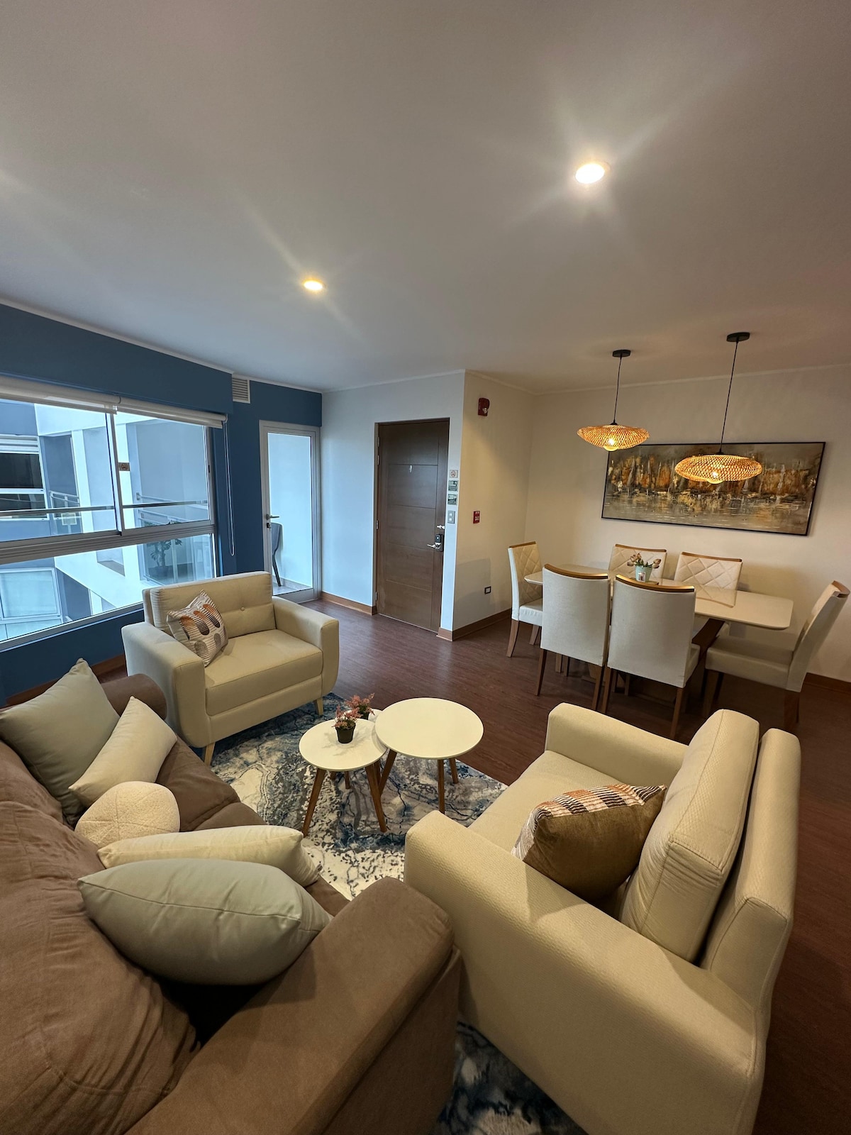 The living area is furnished with two comfortable armchairs and a plush sofa, arranged around a round coffee table. A textured area rug provides warmth to the space. A dining table sits adjacent, illuminated by modern pendant lights, while large windows allow natural light to fill the room.