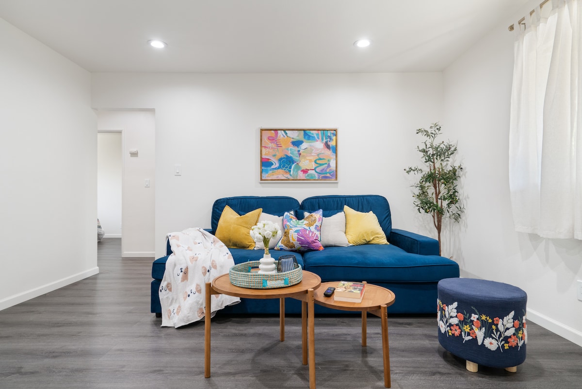 A bright living room features a blue sofa adorned with decorative pillows and a cozy throw. A circular ottoman with floral design complements a wooden coffee table that holds a vase with fresh flowers. Natural light enters through sheer curtains, enhancing the welcoming ambiance.