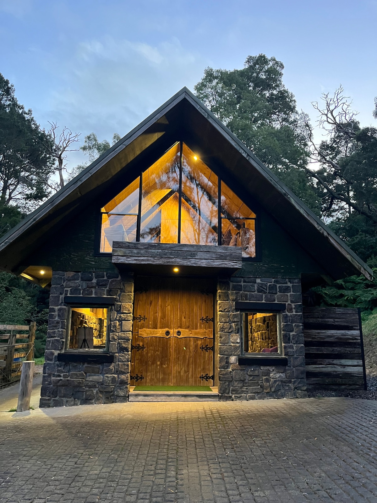 The entrance of a restored bluestone barn is showcased, featuring a striking triangular glass window that reflects the evening sky. Rustic wooden doors, framed by decorative iron accents, are flanked by smaller windows. Surrounded by lush greenery, the pathway leads up to the inviting entrance.
