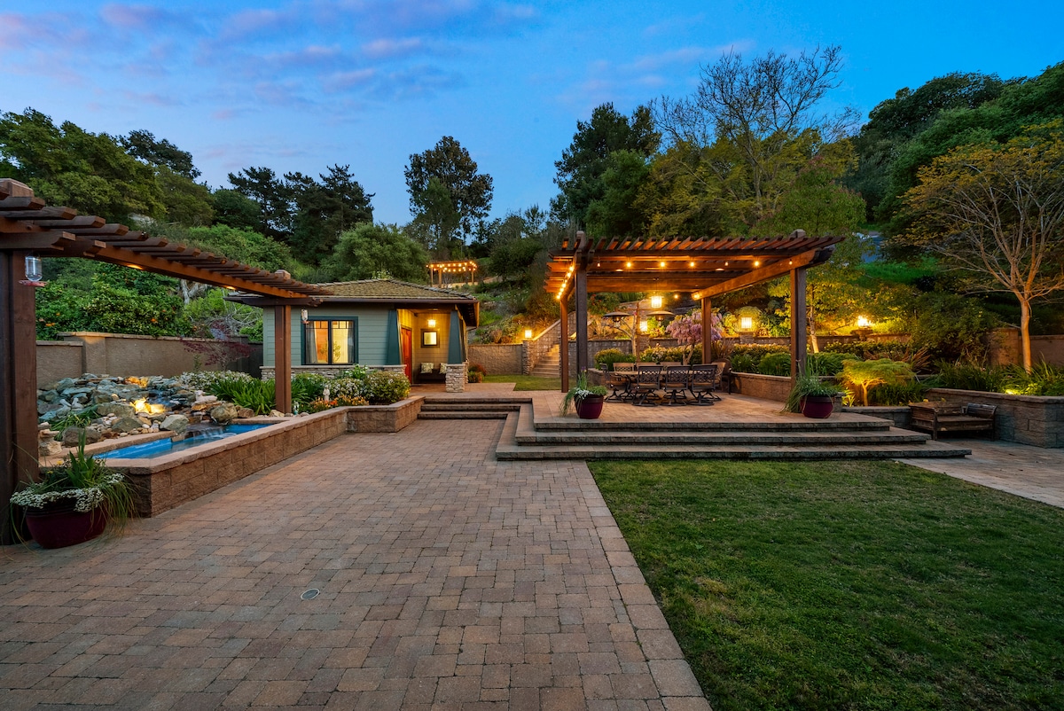 A well-maintained outdoor space features a stone patio illuminated by soft lighting. Multiple seating areas are visible under pergolas, surrounded by lush greenery and flowering plants. A small water feature adds tranquility, while well-placed trees provide shade and privacy.