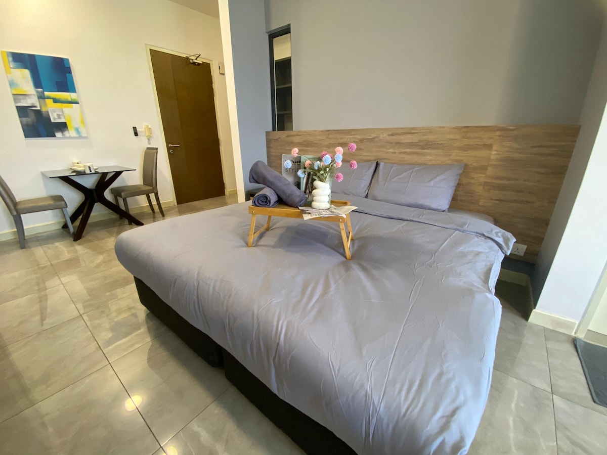 A spacious studio unit features a combined sleeping area with a large bed topped with a gray blanket. A tray with a vase of flowers rests on the bed. In the background, a dining table for two is visible, accompanied by a small artwork on the wall.
