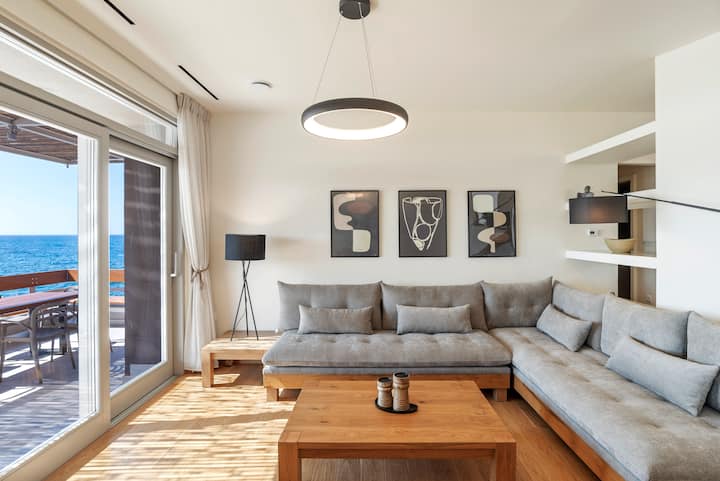 Salon avec 2 canapés d'angle confortables, table basse au milieu, table basse avec lampe, WiFi, fenêtres à gauche, porte coulissante menant à la terrasse donnant sur la mer et mobilier d'extérieur.